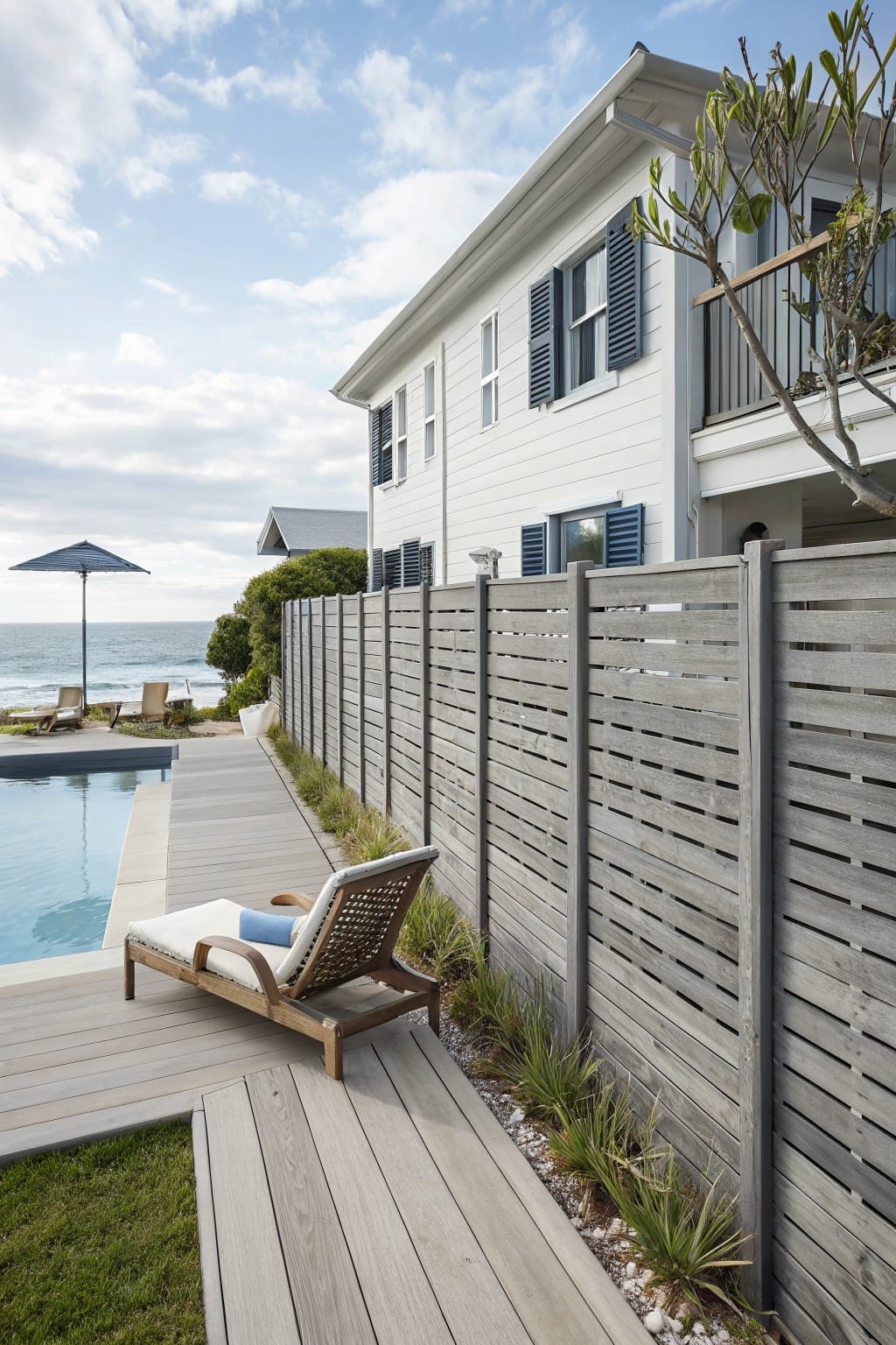 White clapboard house with blue shutters next to an in-ground pool on a wooden deck with lounge chairs, bordered by a tall horizontal slatted gray wood fence and ocean view beyond.