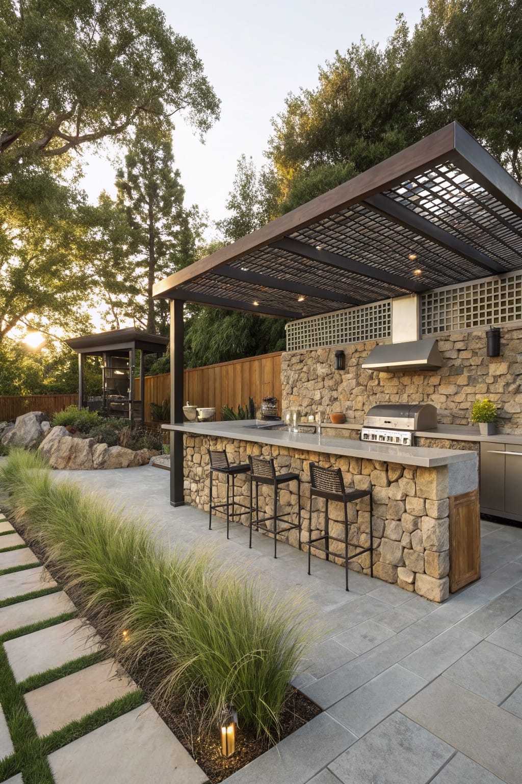 Backyard outdoor kitchen under black metal lattice pergola with tall stone wall backing, black bar stools at stone counter, gas grill, paver pathway, grasses, and trees.