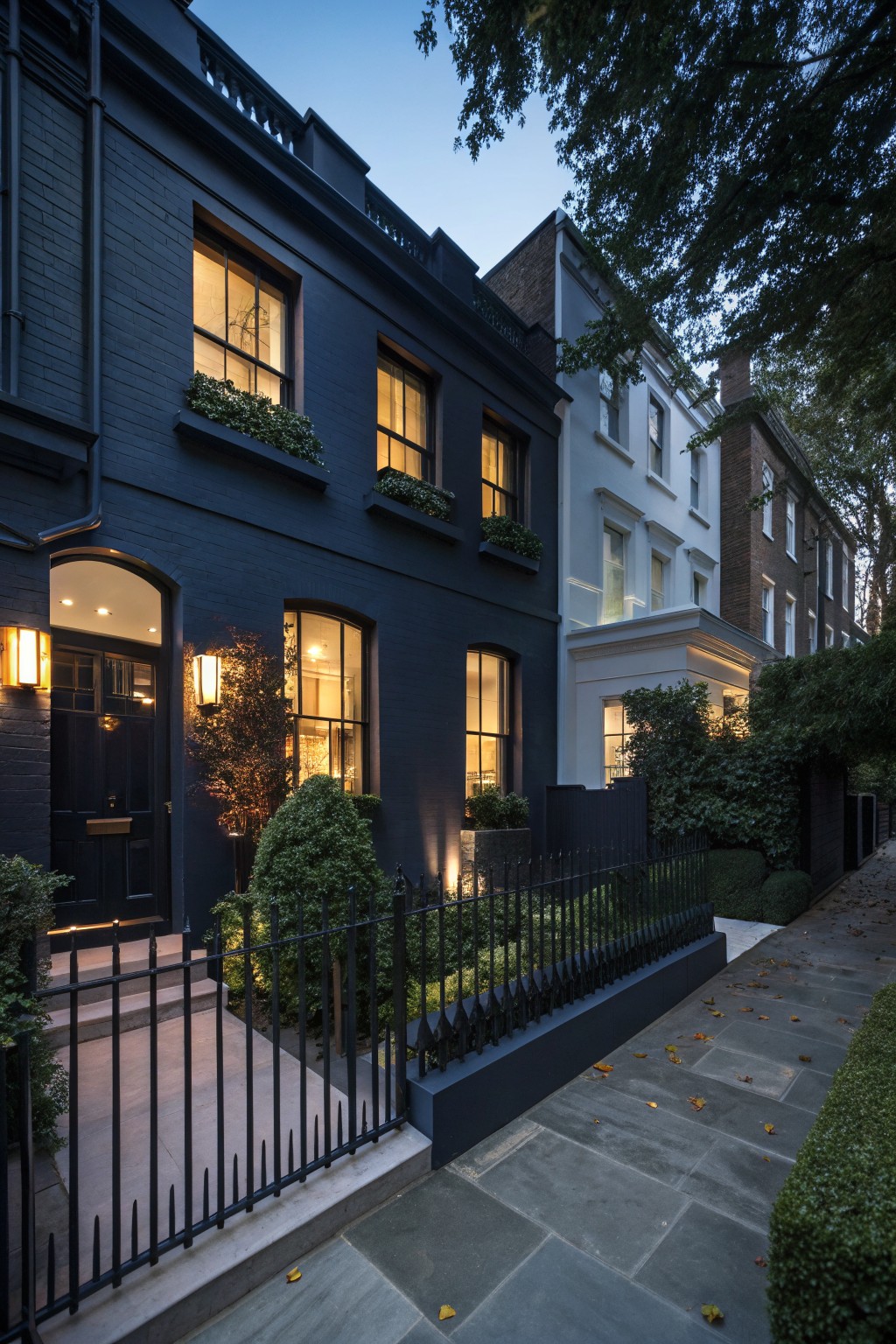 Dark-painted terraced house at dusk with warm lights in windows, low black wrought-iron fence enclosing boxwood topiaries and shrubs, stone steps leading to arched front door.