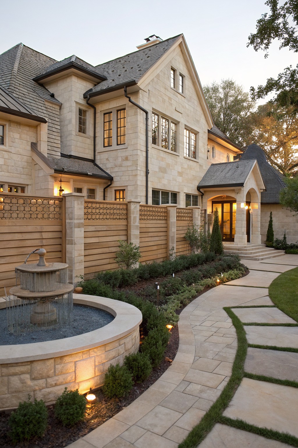 Beige stone house exterior with wooden fence on stone pillars enclosing a front yard garden featuring a circular fountain, curved stone pavers pathway, hedges, and landscape lights at dusk.