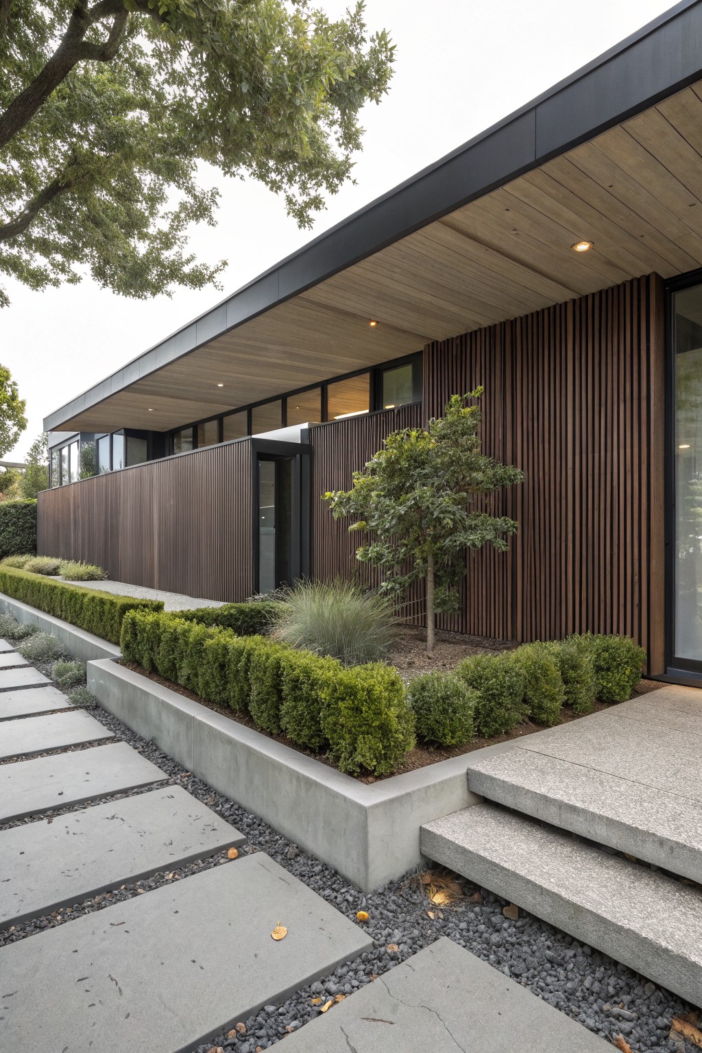 Modern house side exterior with tall vertical dark wood slat wall, raised concrete planter holding boxwood hedges and grasses, concrete paver path and steps to entry door.