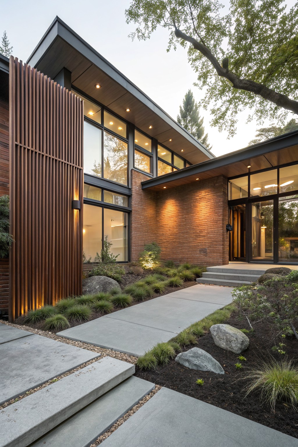 Modern house exterior with tall vertical wood slat screen on brick wall beside concrete pathway and steps, large glass windows, entry door, and front yard landscaping of grasses, boulders, and low plants.