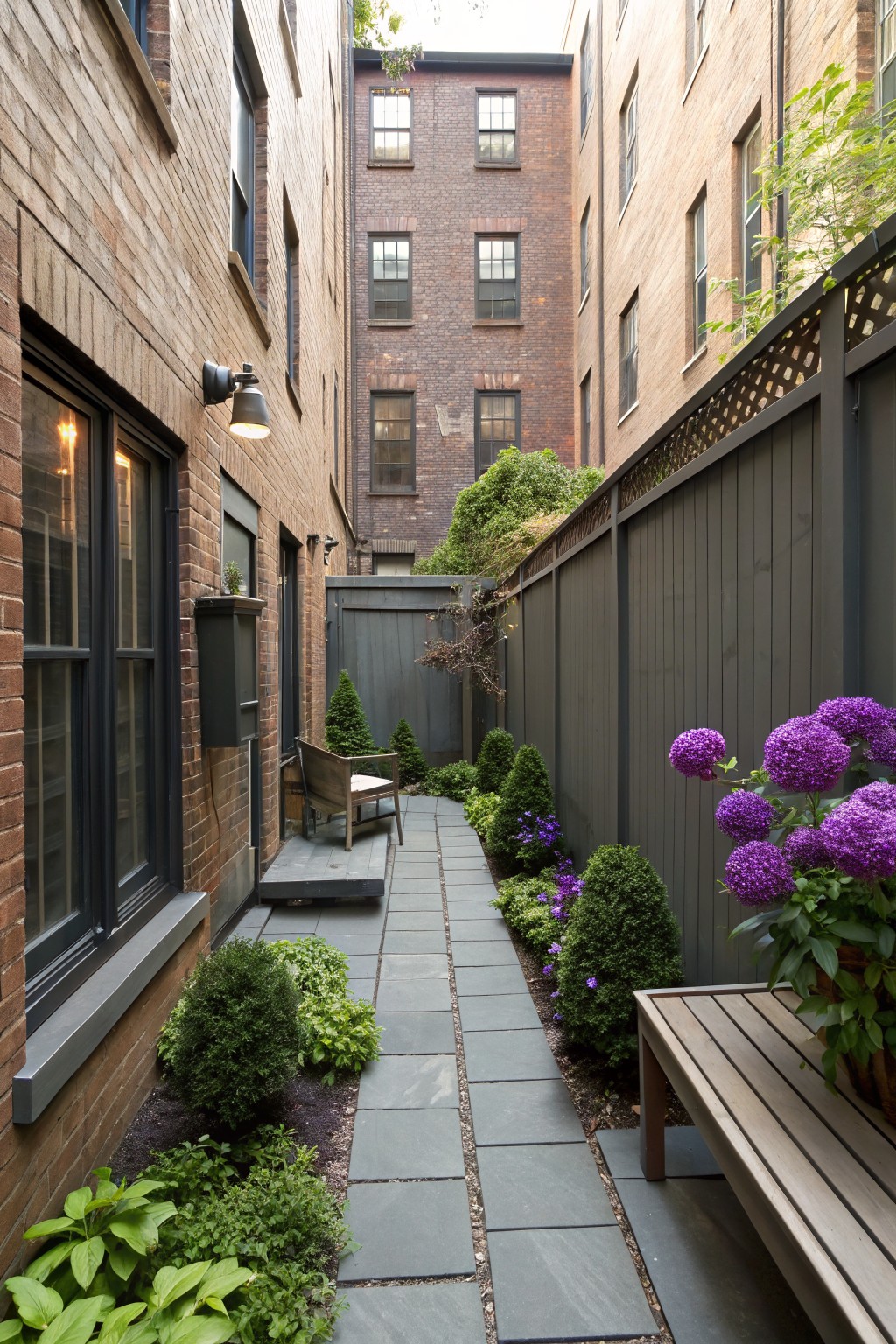Narrow courtyard between brick buildings with gray slate pathway lined by boxwood shrubs, purple blooming flowers, hostas, wooden benches and chair, and gray fence.