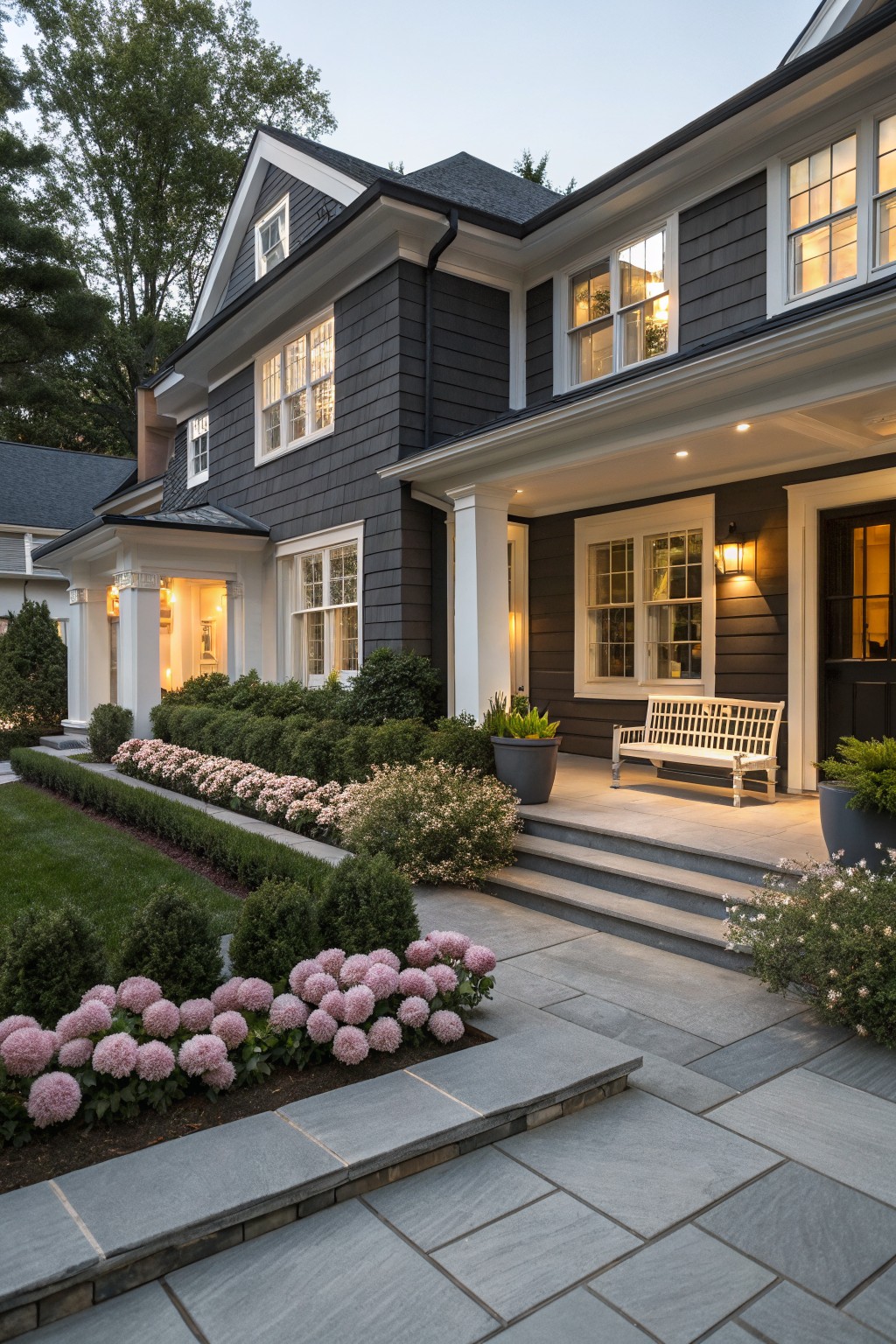 Dark gray shingled house with white trim and covered porch approached by stone steps and walkway bordered by trimmed boxwood hedges and pink blooming hydrangeas.