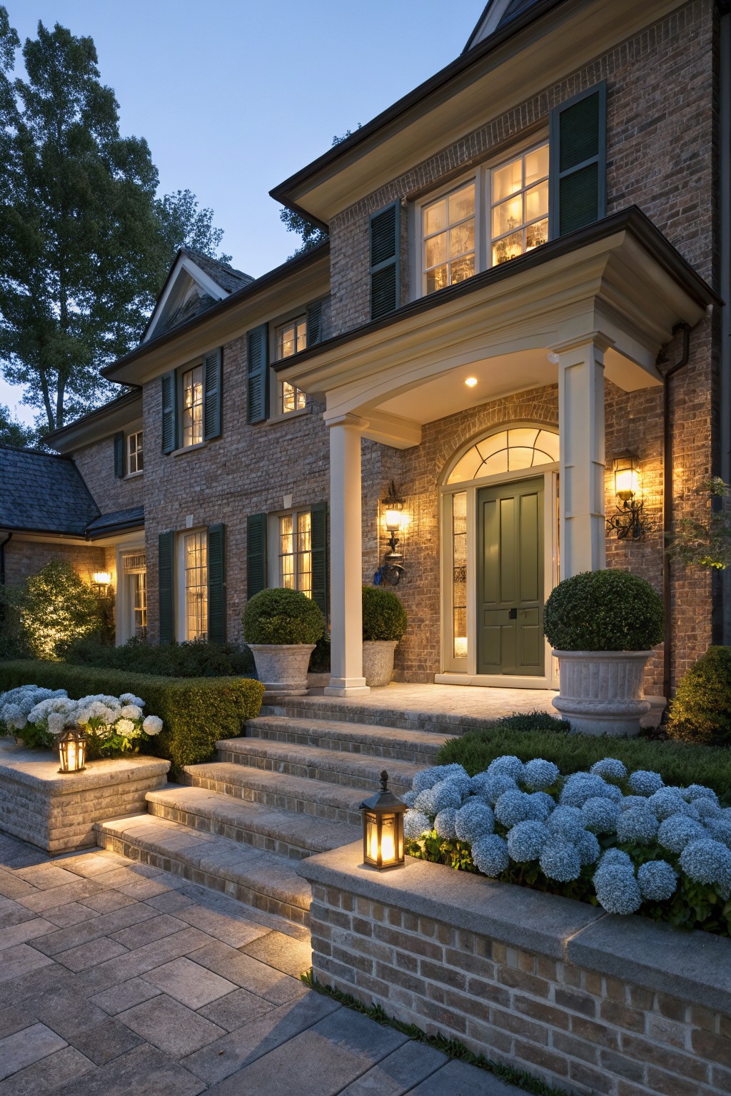 Brick house exterior at dusk with columned portico, green front door, large potted spherical boxwoods flanking the entry landing, hydrangea plantings along steps and walls, stone pathway, and lantern lighting.