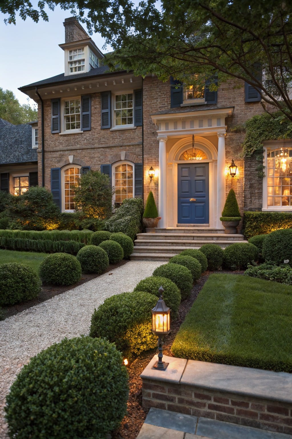 Brick house facade with blue front door under a portico, gravel pathway edged by spherical boxwood shrubs and low hedges, formal landscaping with uplights at dusk.