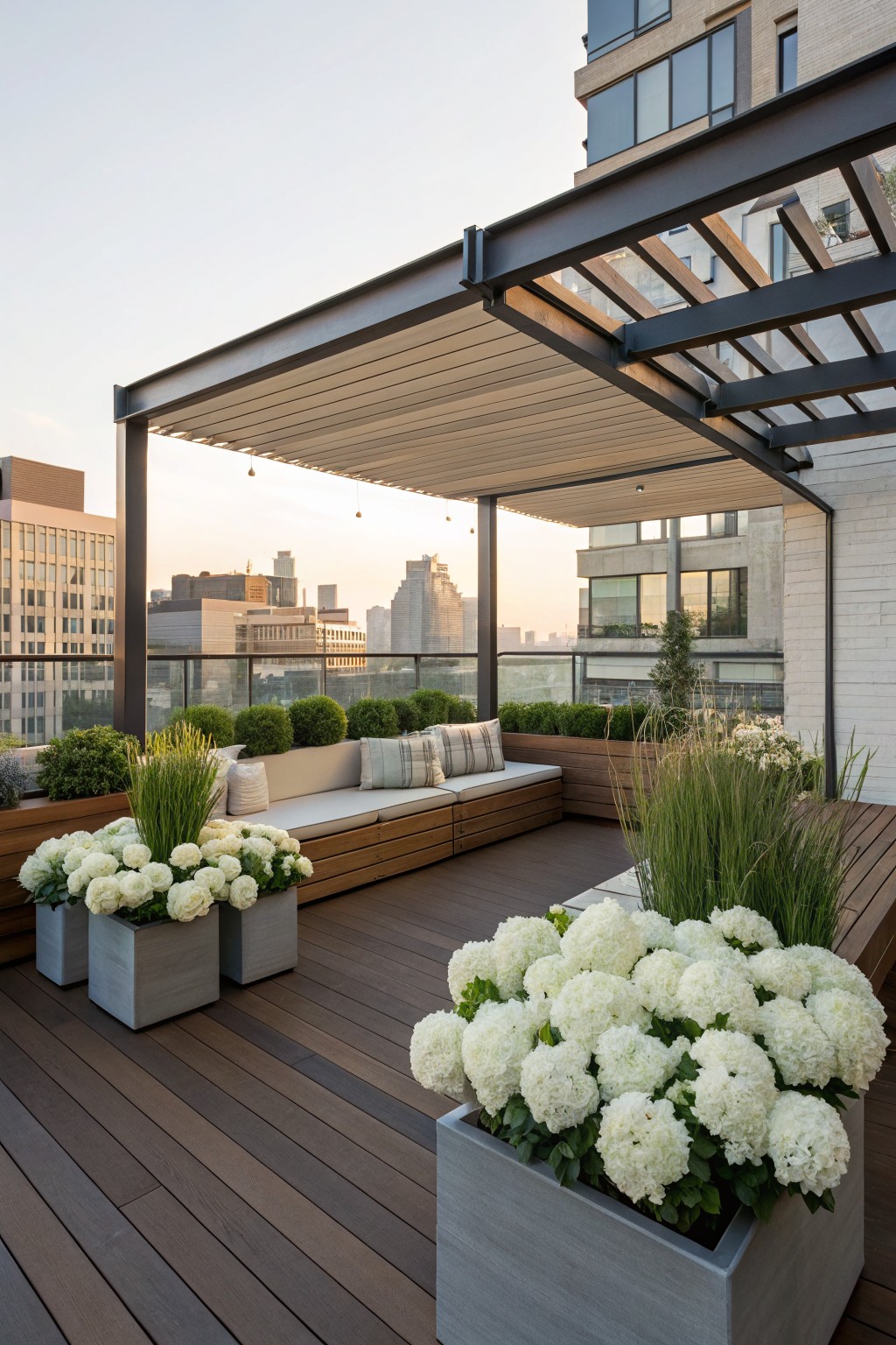 Rooftop terrace with wooden decking, built-in cushioned benches integrated with planter boxes of white hydrangeas and tall grasses, under a black metal pergola with retractable beige shade sail, city skyline in background.