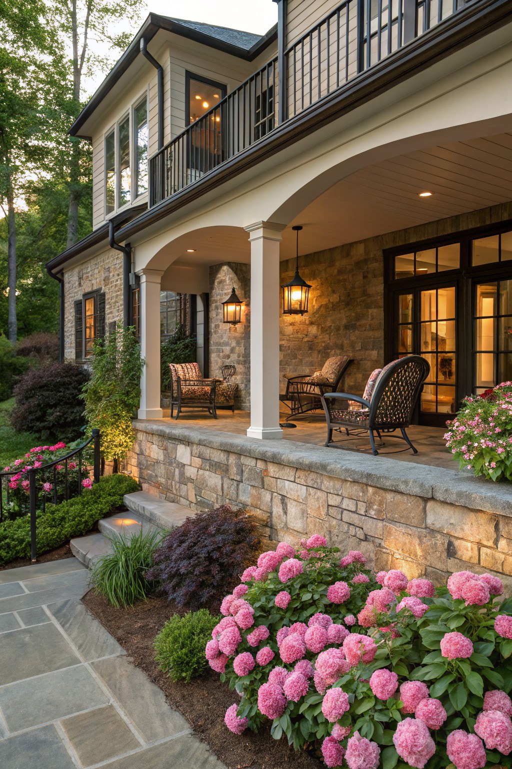 Stone house porch with wicker chairs under arches, steps and bluestone path leading down through beds of pink mophead flowers, shrubs, and trees in evening light.