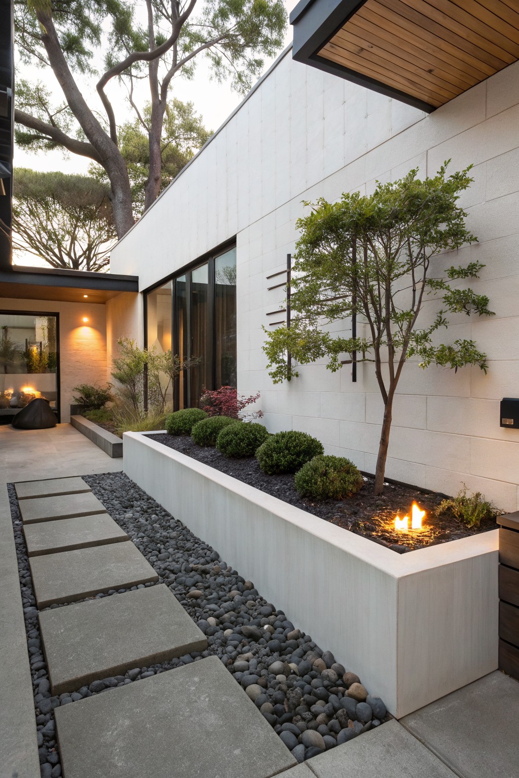 Long white raised concrete planter bed along a gravel and concrete stepping stone path, with embedded yellow gas fire flames amid black mulch and low green shrubs, next to a white stucco house wall with windows and trees.