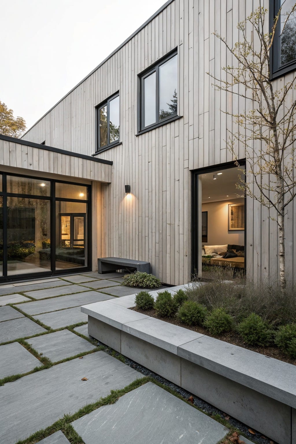 Modern house corner with light vertical wood siding, black-framed windows and glass doors opening to a gray slab patio edged by a concrete bench, low shrubs, grasses, and a young tree.