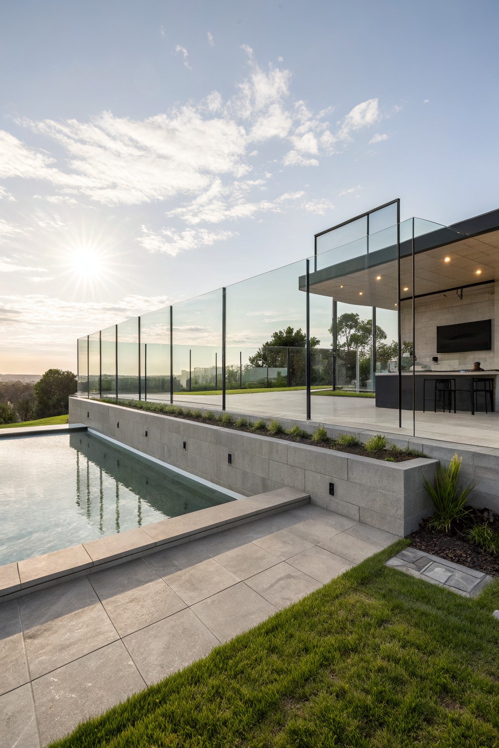 Backyard infinity pool with frameless glass balustrade fencing along the edge and deck, concrete retaining walls with plants, outdoor kitchen with black cabinetry and bar stools, and surrounding grass and trees at sunset.
