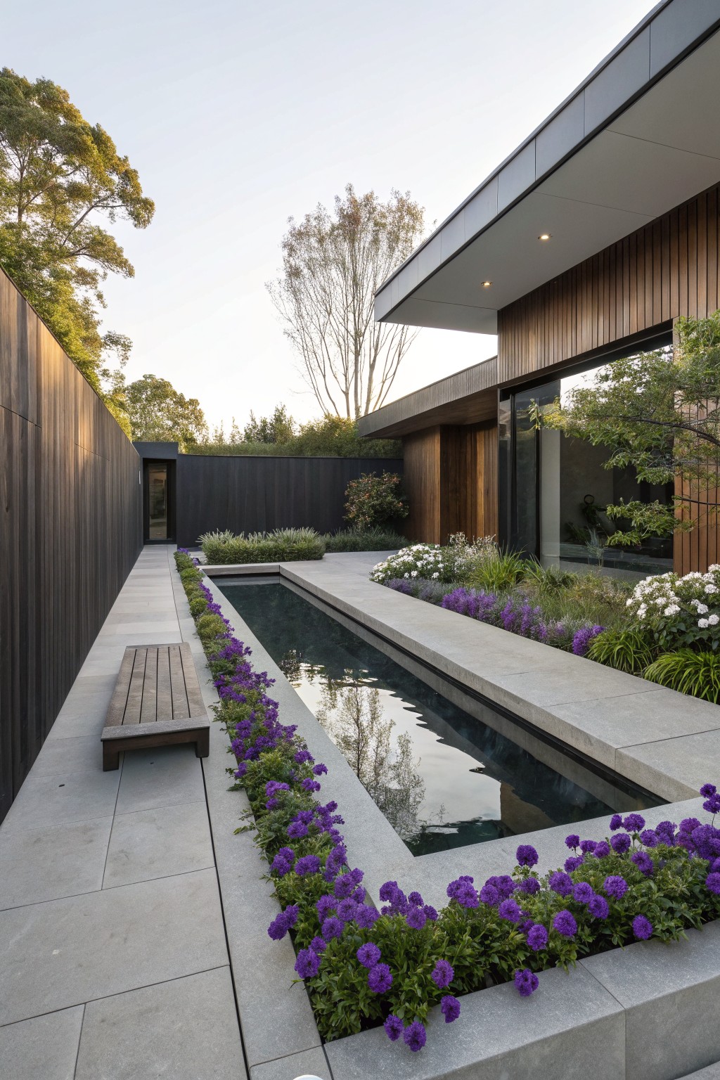 Narrow rectangular reflecting pool bordered by purple ageratum flowers on one side and a wooden bench on a stone path, with dark wooden fencing, trees, and a modern wood-clad house visible.