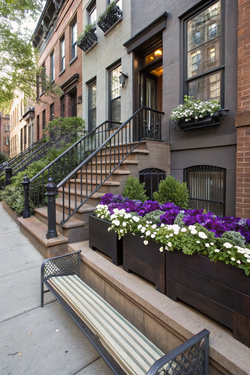 City brownstone with black wrought-iron stairs to the entrance, large wooden planters at the base overflowing with purple and white pansies and other flowers, plus a striped bench on the sidewalk.