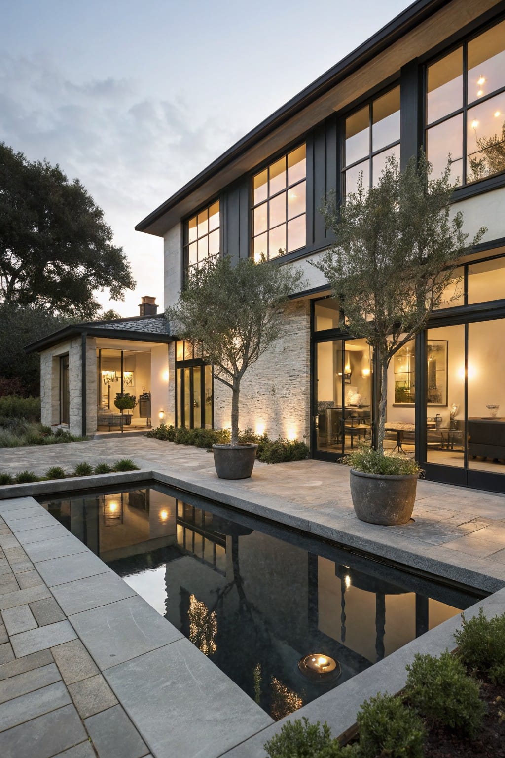 Modern two-story house exterior at dusk with large black-framed windows, a rectangular black reflecting pool in the stone-paved backyard patio flanked by olive trees and potted plants.