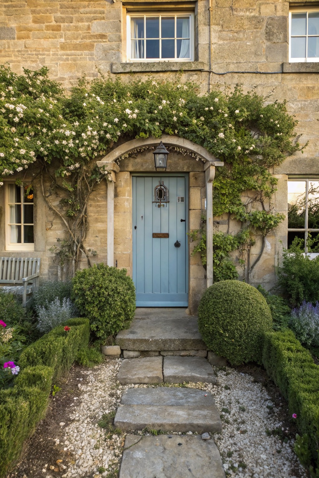 Stone cottage with arched porch entry covered in white-flowering climbing vines over a blue front door, stone steps, boxwood shrubs, and gravel path.