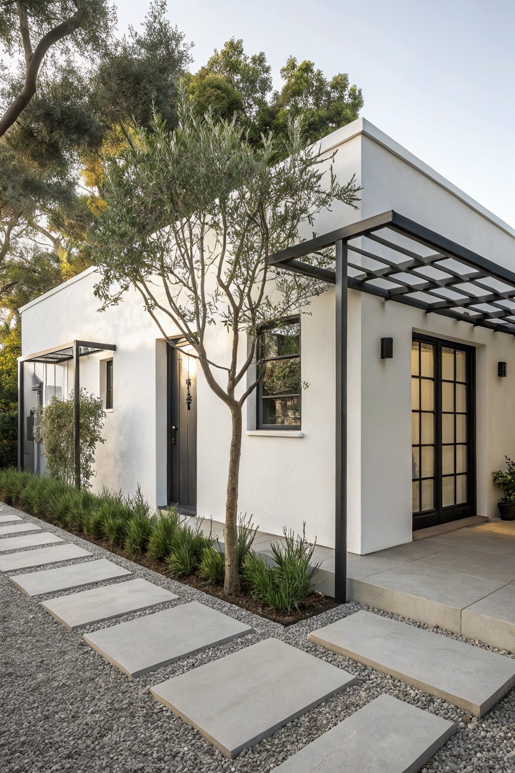 White stucco house exterior featuring a black metal pergola over the front door and patio, with a young olive tree nearby, concrete paver stepping stones set in gravel, and low green plantings along the edge.