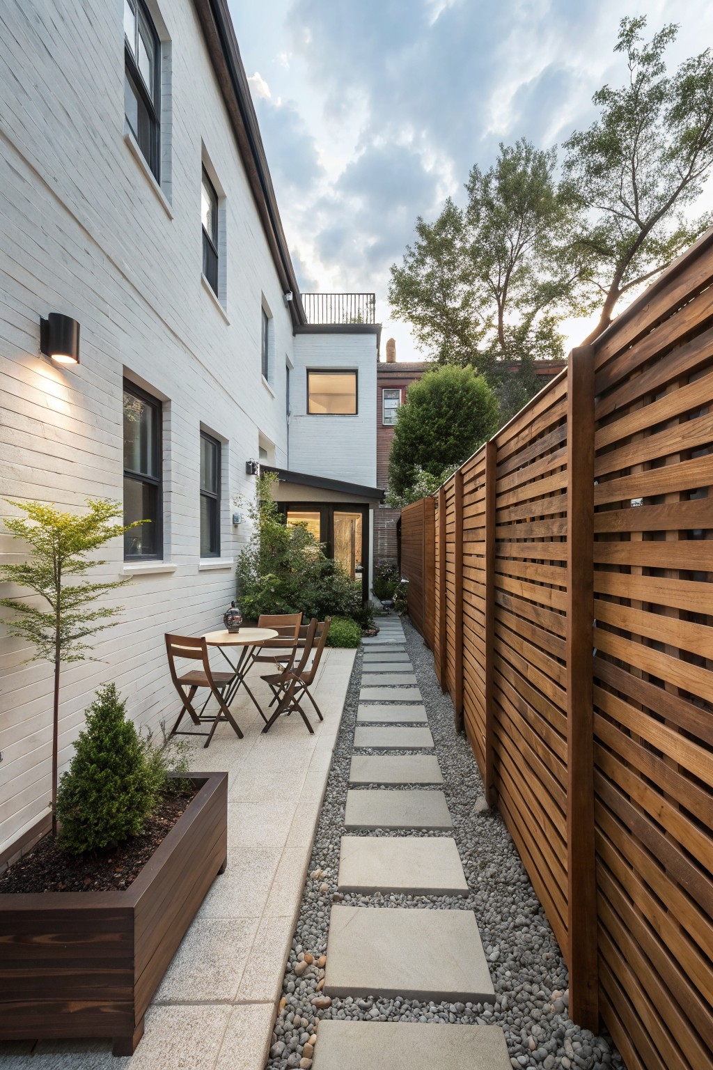 Narrow outdoor pathway lined with gravel and stepping stones between a white brick wall on one side and tall horizontal wooden slat fence on the other, featuring potted plants, a small table with chairs, and greenery.