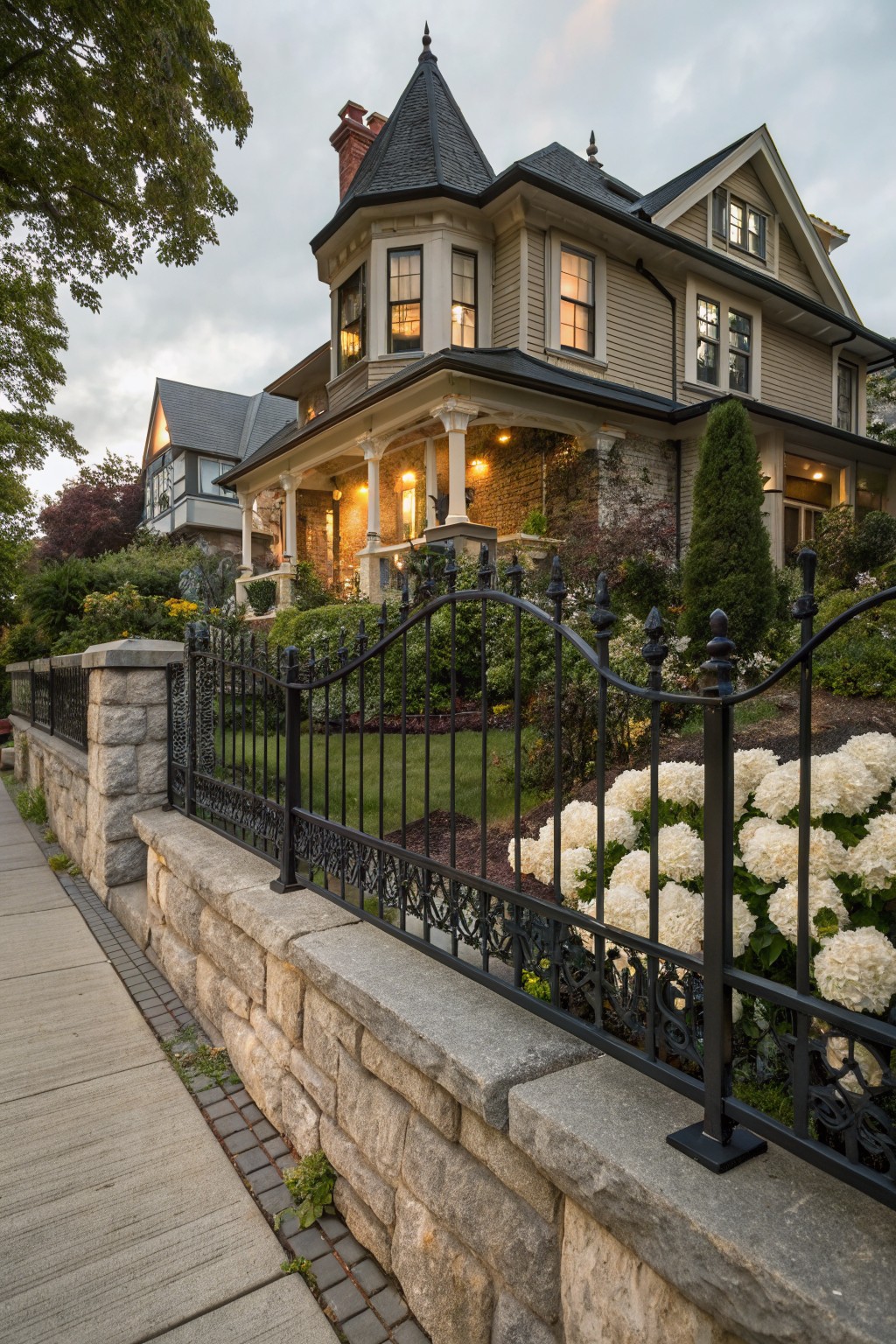 Beige Victorian house with turret and porch behind curved black wrought iron fence on stone retaining wall, white hydrangeas and landscaping along sidewalk at dusk.