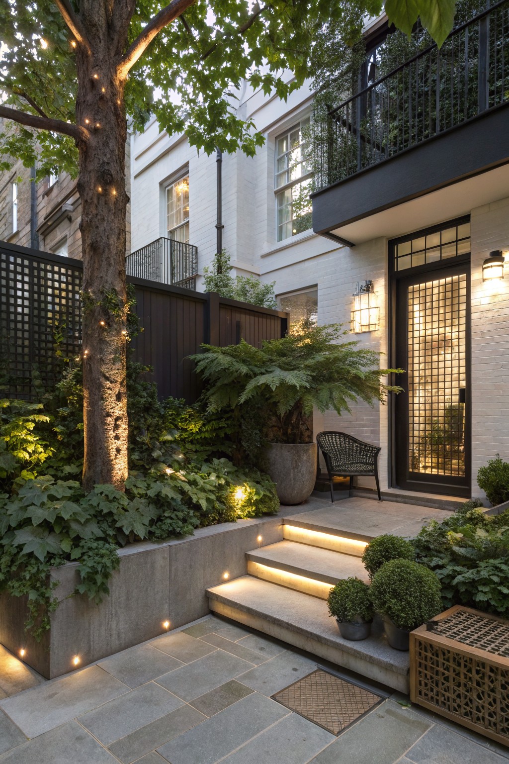 Garden path with concrete steps lit from below leading to a black metal door on white brick wall, large tree trunk wrapped in string lights providing overhead shade, surrounded by ferns, potted plants, and an outdoor chair.