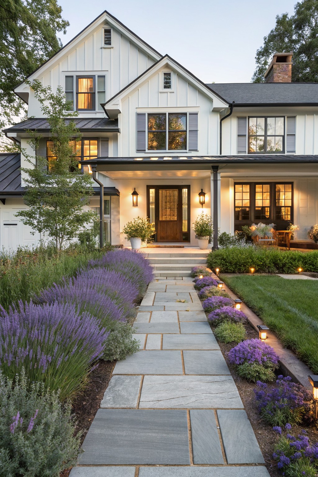 White gabled house with black metal porch roof and trim, gray stone walkway lined with lavender plants and small path lights leading to wood front door, flanked by lawn, shrubs, and potted plants.