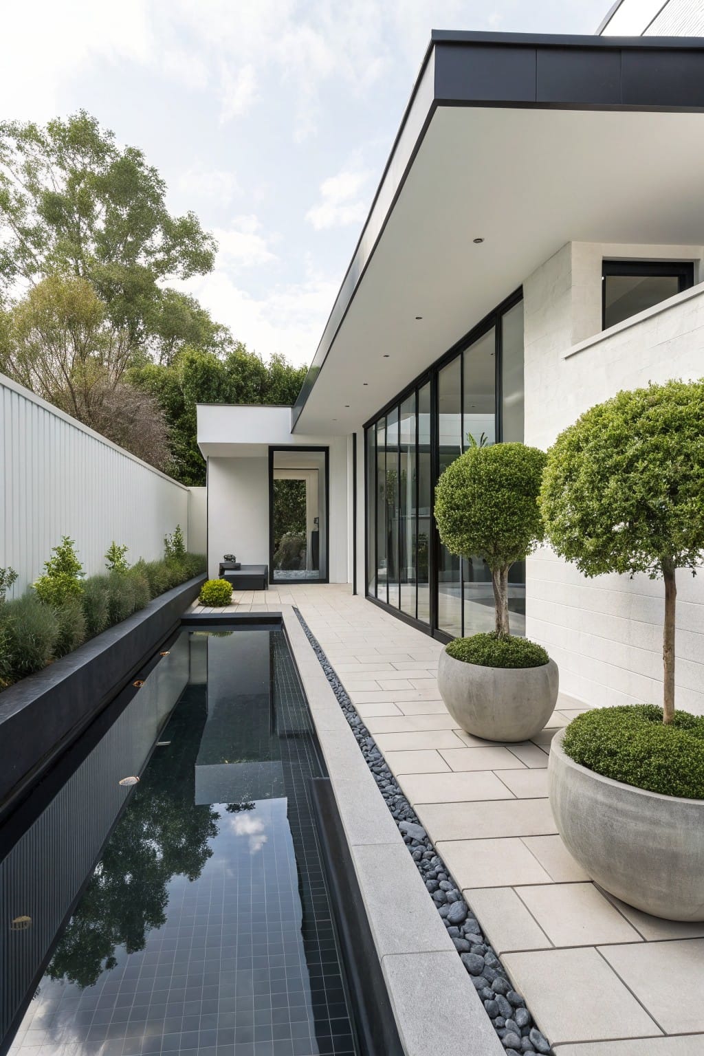 Long narrow reflecting pool with black tiles runs parallel to a white modern house exterior featuring black-framed glass walls and doors, bordered by pebbles and a light stone pathway with potted plants nearby.