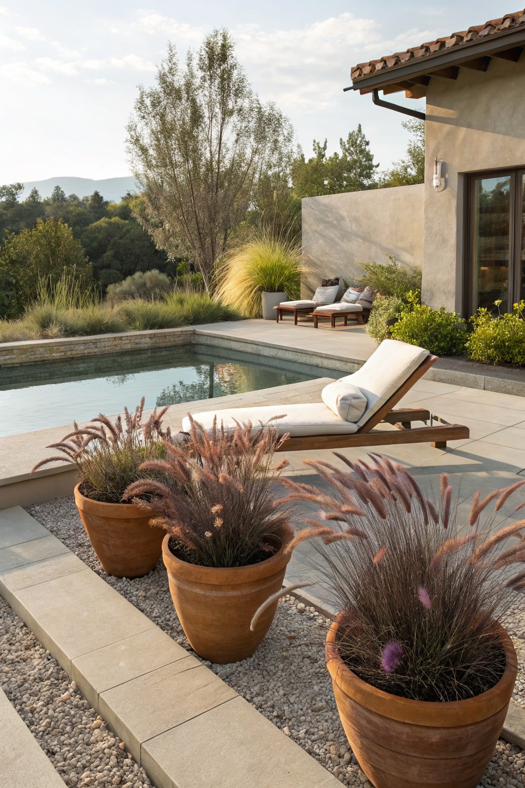 Pool deck with white lounge chair and pillow next to three large terracotta pots of tall pinkish ornamental grasses, beside a rectangular pool and stucco house wall with trees and hills in background.