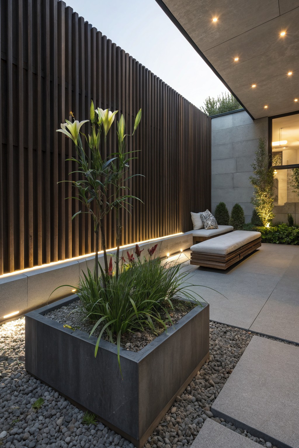 Modern outdoor patio with dark vertical slat wood fence, tall white daylilies planted in front, square concrete planter with grasses, wooden bench on pavers, concrete walls with LED lighting strips, and small gravel areas.