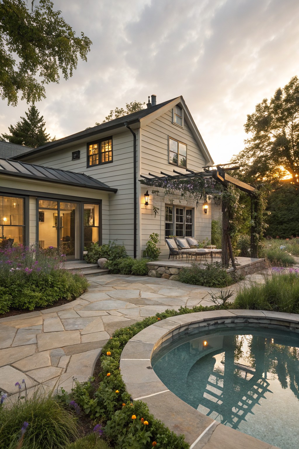 Gray shingle house with gabled roof and glass doors opening to a pergola-covered patio with chairs, a curved flagstone path leading through plants to a round in-ground hot tub, surrounded by grasses and trees at dusk.