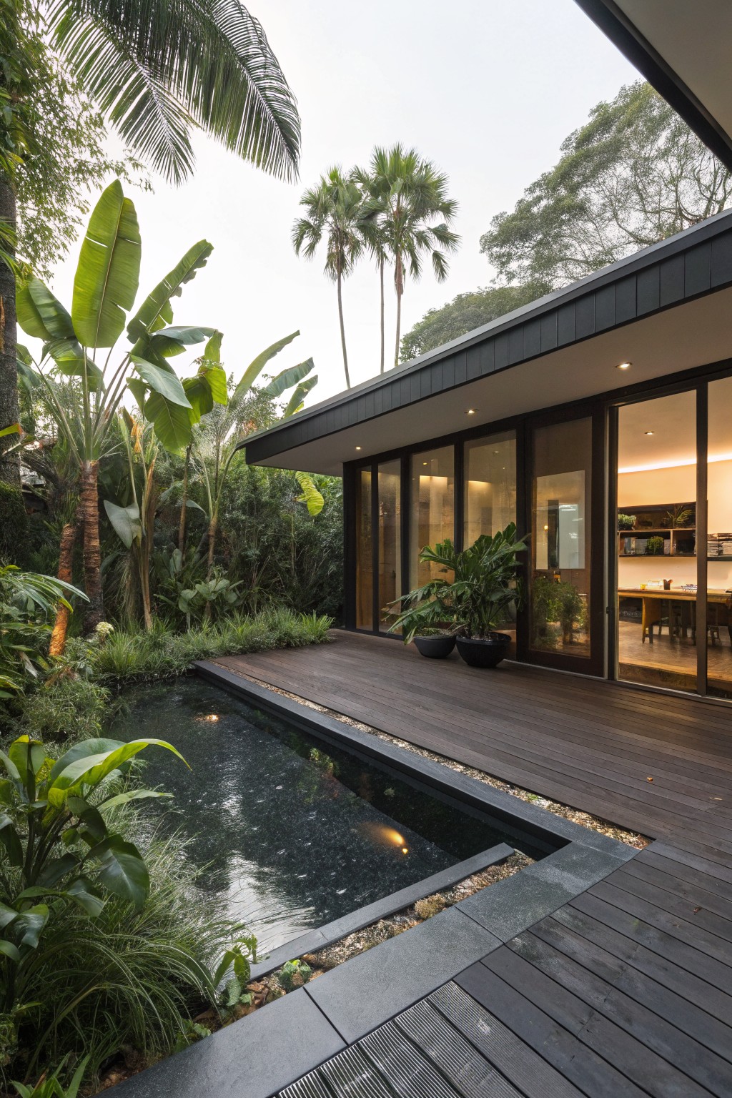 Modern backyard with dark wooden deck adjacent to narrow black pool, surrounded by tropical plants like palms and bananas, and glass walls of house visible.