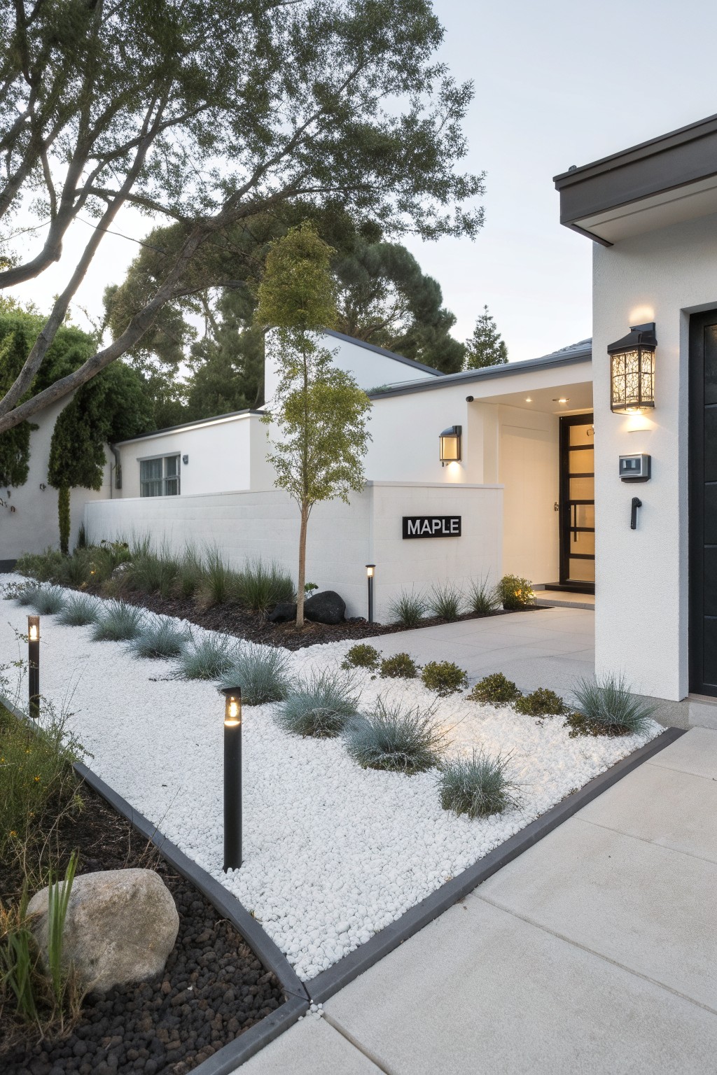 Modern white stucco house exterior with black garage door and entryway, featuring landscaping of white pebble beds, blue ornamental grasses, black mulch borders, larger rocks, and black pathway lights leading to the front door.