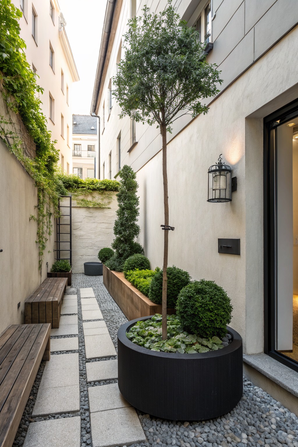 Narrow passageway between light-colored buildings featuring a central slender tree surrounded by spherical boxwood shrubs, raised wooden planters with greenery, a pebble and stone path, wooden bench, and wall lantern.
