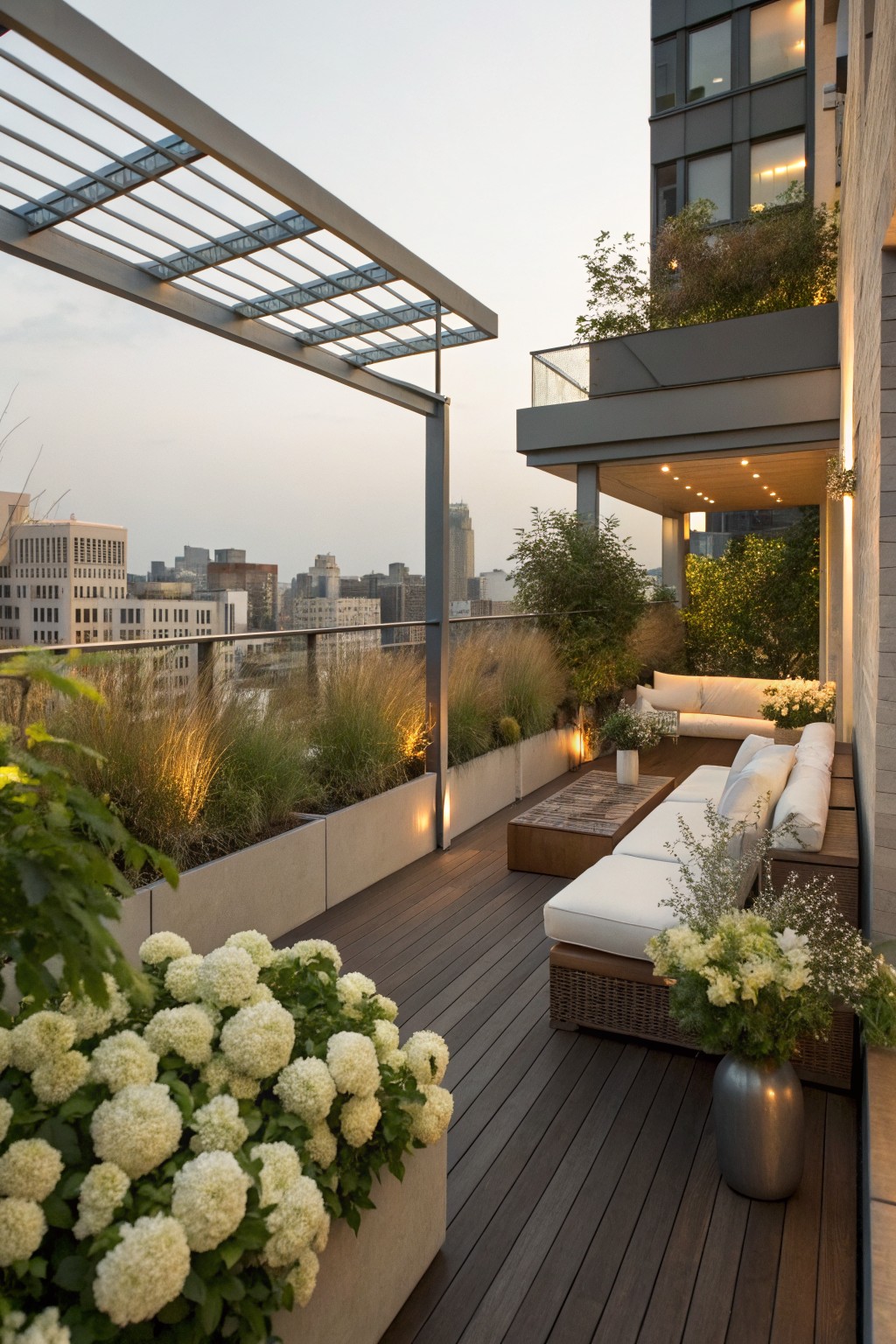 Rooftop terrace balcony with white upholstered sofas and ottomans around a wood coffee table, bordered by large concrete planters filled with white hydrangea bushes and ornamental grasses, overlooking city skyline at dusk.