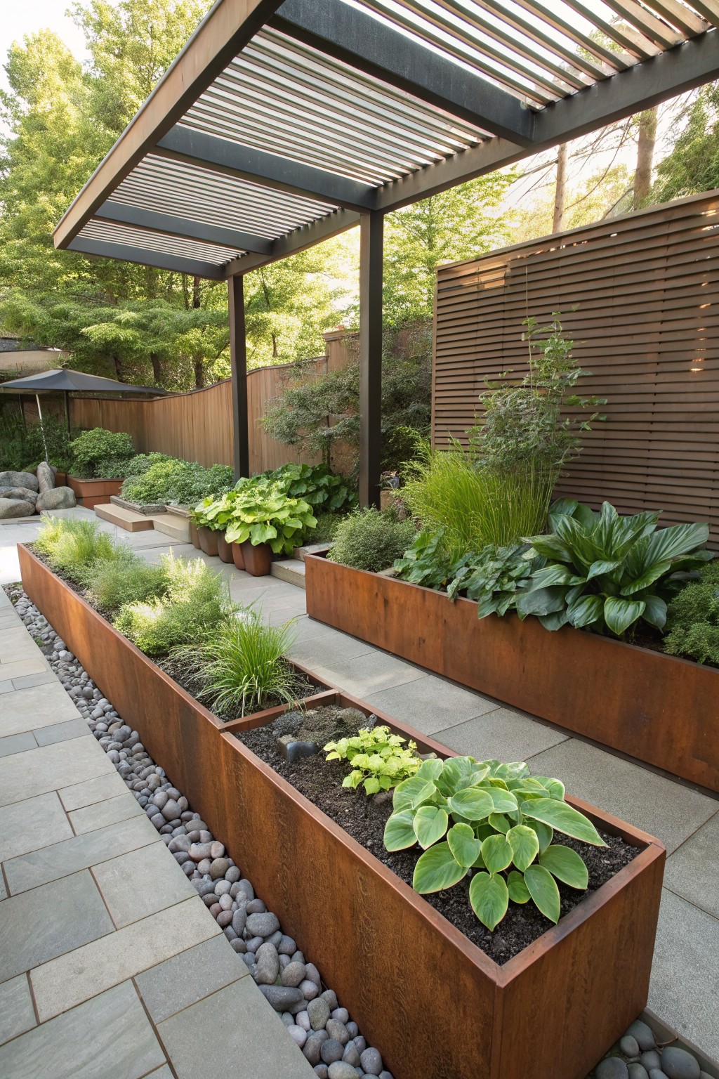 Long rectangular corten steel planters filled with hostas, grasses, and leafy plants line a gray stone paver path edged with pebbles in a backyard garden under a slatted wooden pergola.