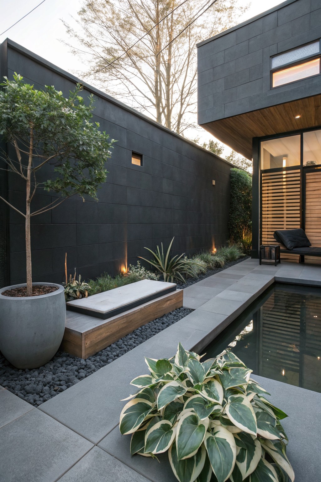 Gray slate paver path beside a narrow pool, edged with a large variegated hosta plant, black gravel mulch, and a wooden bench on a deck against a dark brick wall.