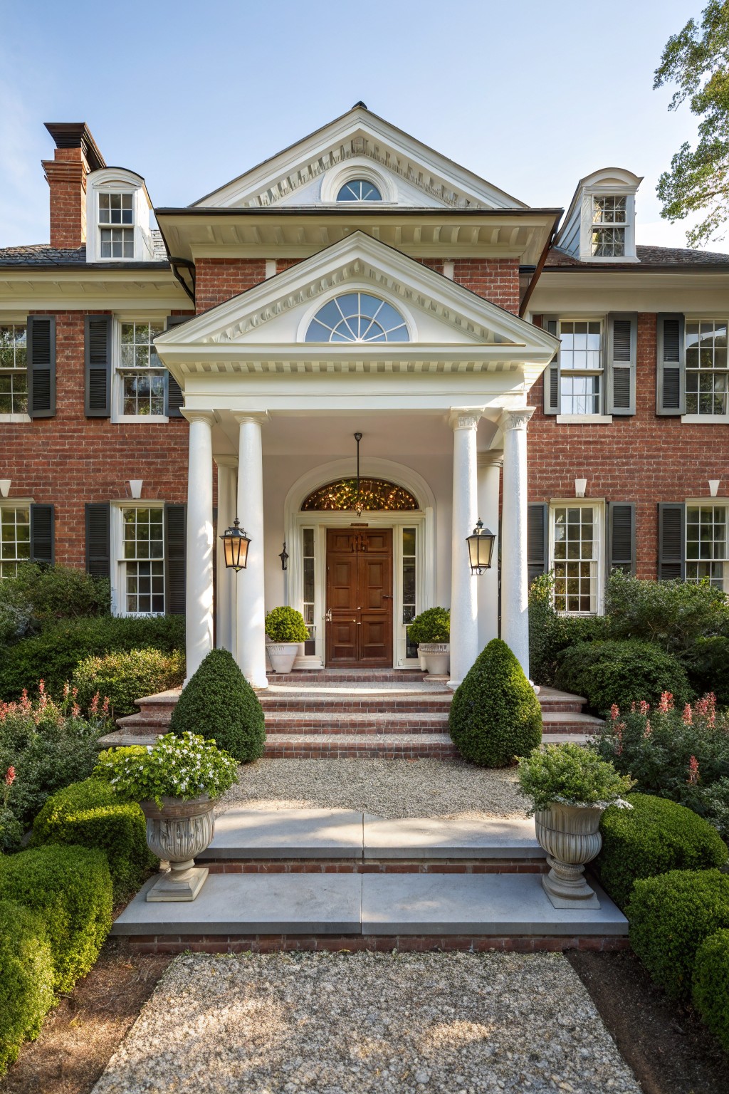 Red brick house with white columns and pedimented porch, symmetrical gravel path lined with boxwood topiaries in urns and pots, flower beds with green shrubs and red blooms leading to front steps.