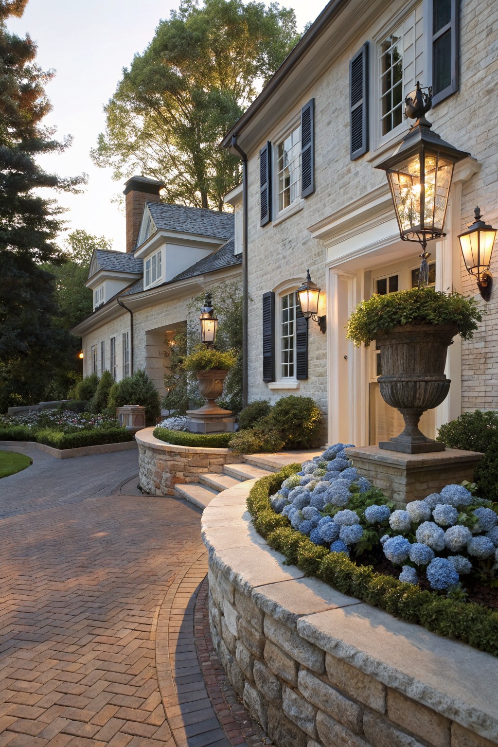 Light beige brick house facade with black shutters and white trim, curved brick paver pathway leading to front door, low stone retaining wall planted thickly with blue hydrangea blooms, flanked by lanterns and topiary planters.