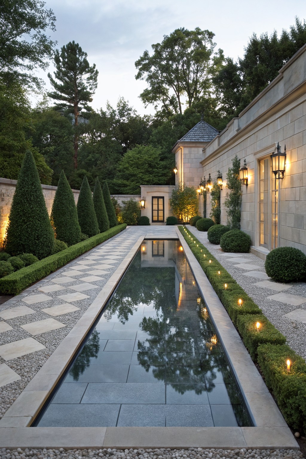Narrow black reflecting pool centered in a formal garden pathway of light and dark checkered stone pavers edged with white gravel and boxwood hedges, with stone walls, trees, and lanterns on both sides.