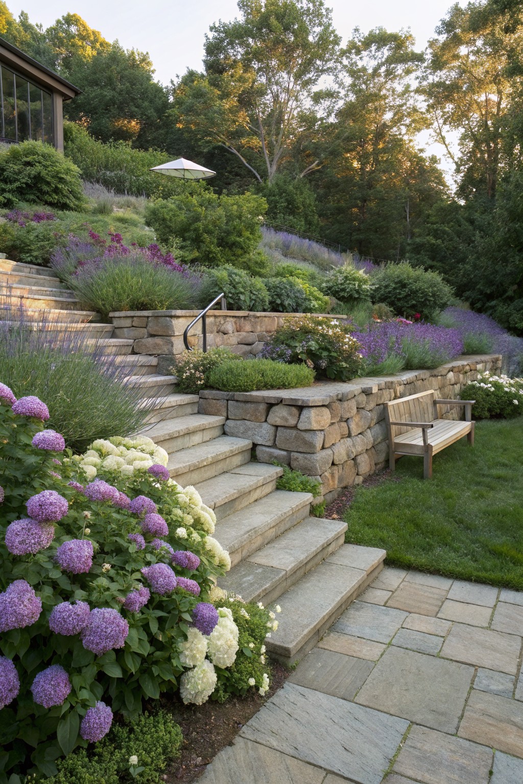 Stone steps on a sloped garden site edged with purple and white hydrangea blooms, lavender plants, shrubs, and stone retaining walls, with a wooden bench nearby and trees and a house in the background.
