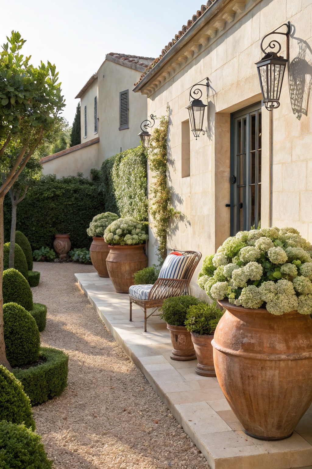 Beige stone house exterior wall with arched door, wall lanterns, large terracotta pots of white hydrangeas, boxwood topiaries, gravel path, and wicker chair on raised stone platform.