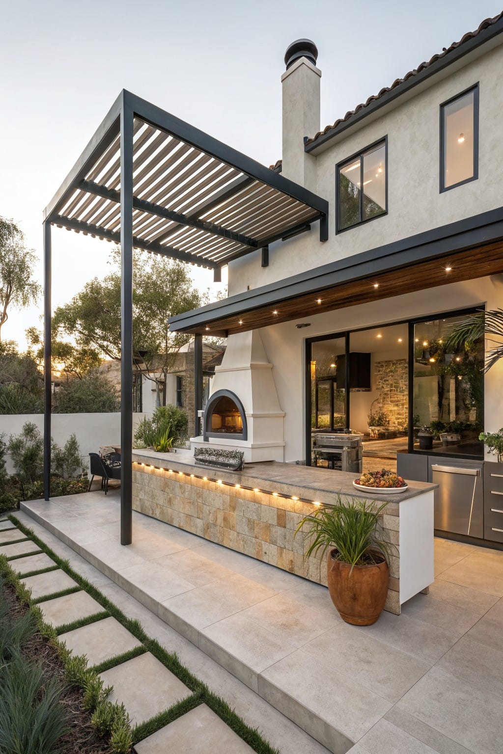 Backyard patio with built-in outdoor kitchen island featuring wood-fired pizza oven, under black slatted pergola, next to white stucco house with glass doors, stone pathway, potted plants, and low landscaping.
