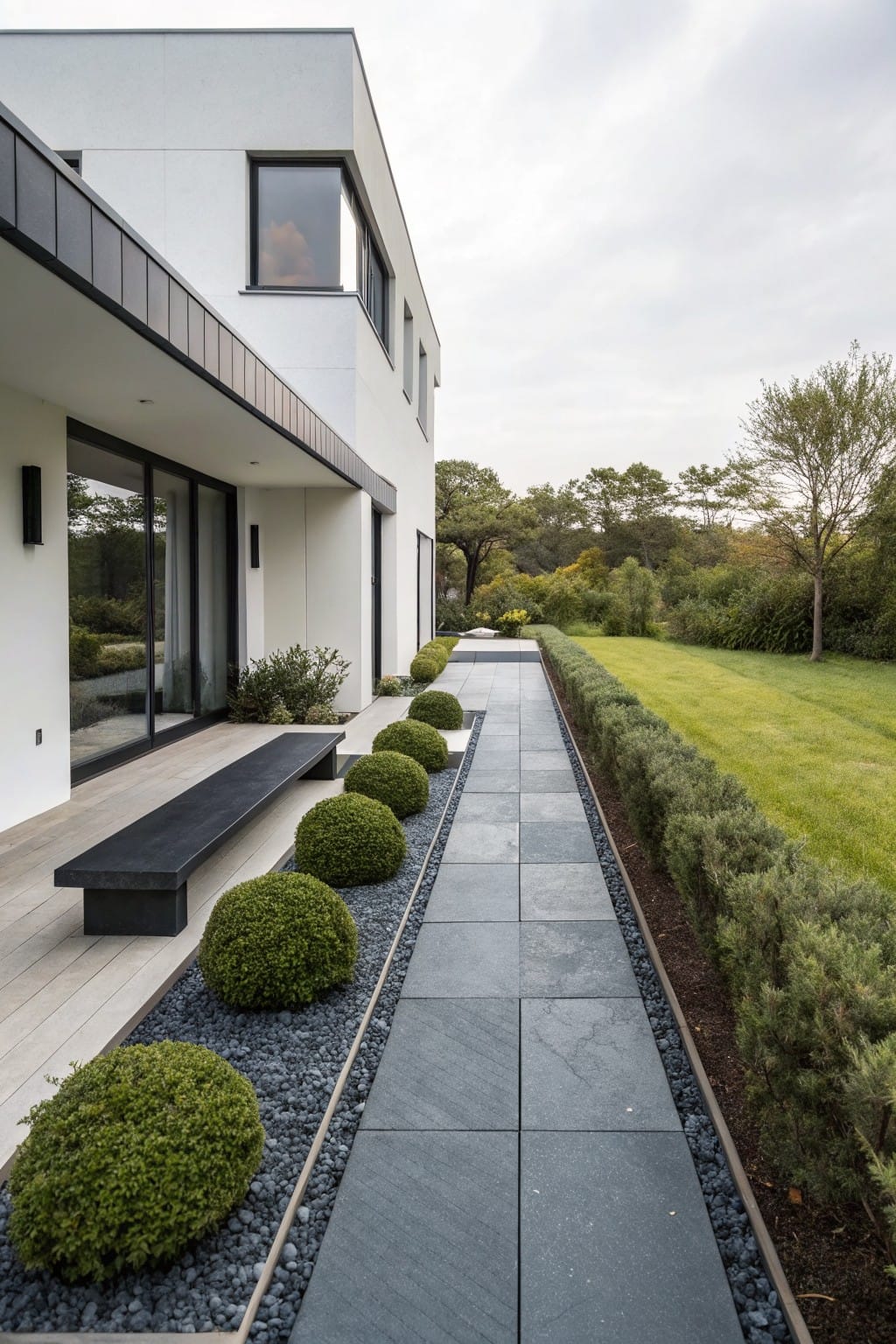 White modern house with wooden deck and black bench beside a dark slate tile pathway edged by gravel strip, boxwood spheres, and grass lawn bordered by hedges.