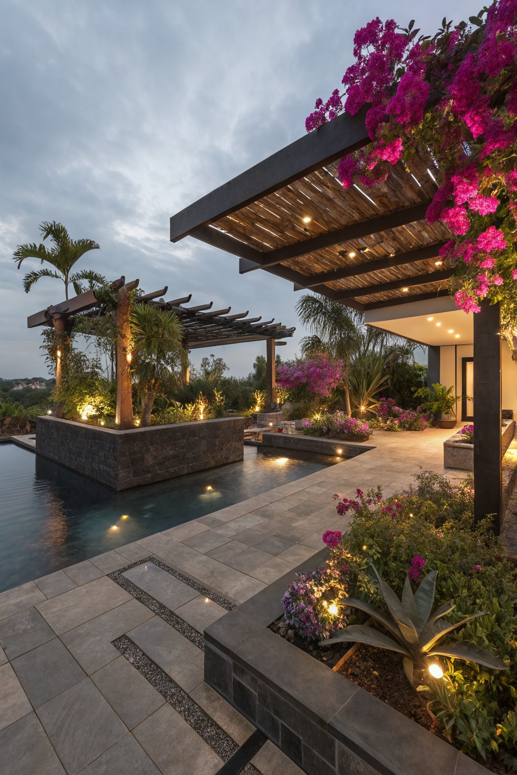 Dusk view of an infinity-style pool in a raised black stone surround on a stone terrace deck, framed by wooden pergolas covered in pink bougainvillea vines, tropical plants, palms, uplights, and nearby modern house exterior.