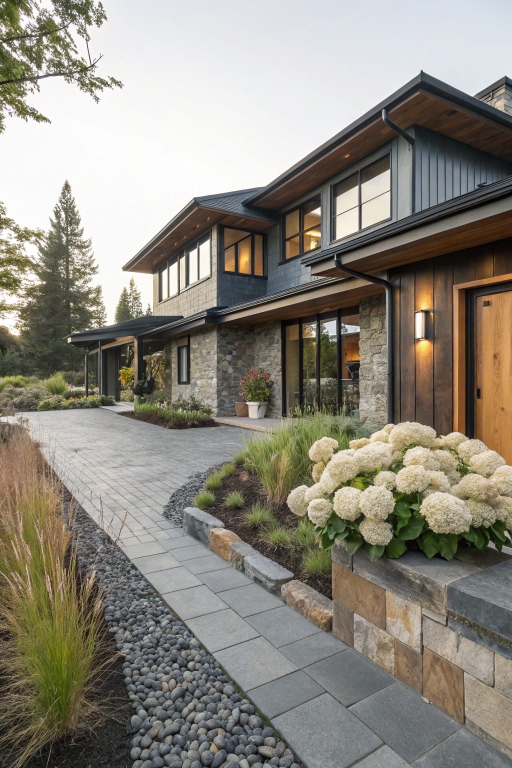 Side exterior of a two-story modern house with dark siding and stone base, featuring low stone retaining walls edging gravel beds planted with hydrangeas and ornamental grasses beside a gray paver pathway leading to a wooden entry door.