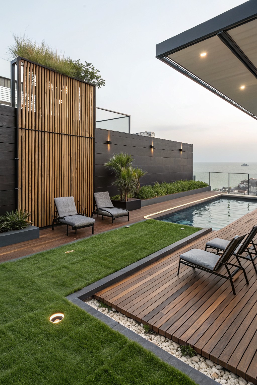 Rooftop terrace with wooden decking, infinity-edge pool, artificial grass lawn strip, lounge chairs, potted plants, wooden slat screens, and dark walls overlooking ocean at dusk.