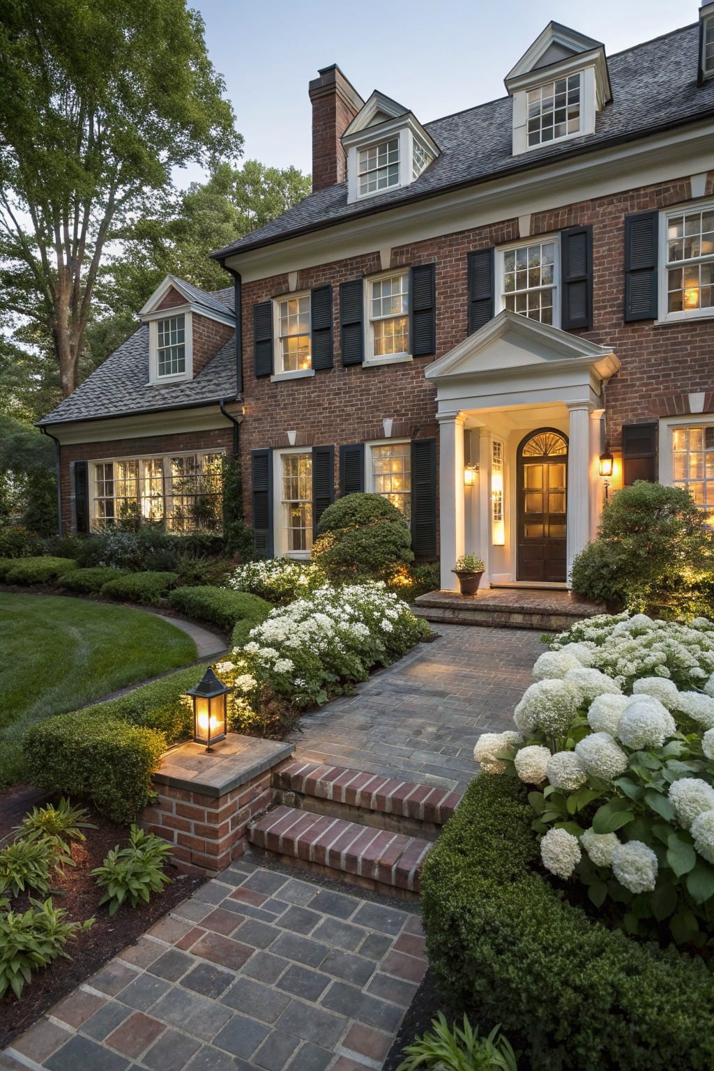 Brick pathway edged with boxwood shrubs and white hydrangea blooms leading to the columned entry of a brick house at dusk, with lanterns providing warm light.