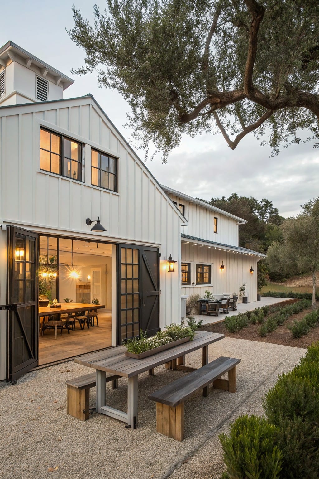 White barn-style house with large open black garage doors revealing an interior dining table, next to a gravel patio holding a wooden picnic table and benches, edged by shrubs and olive trees.