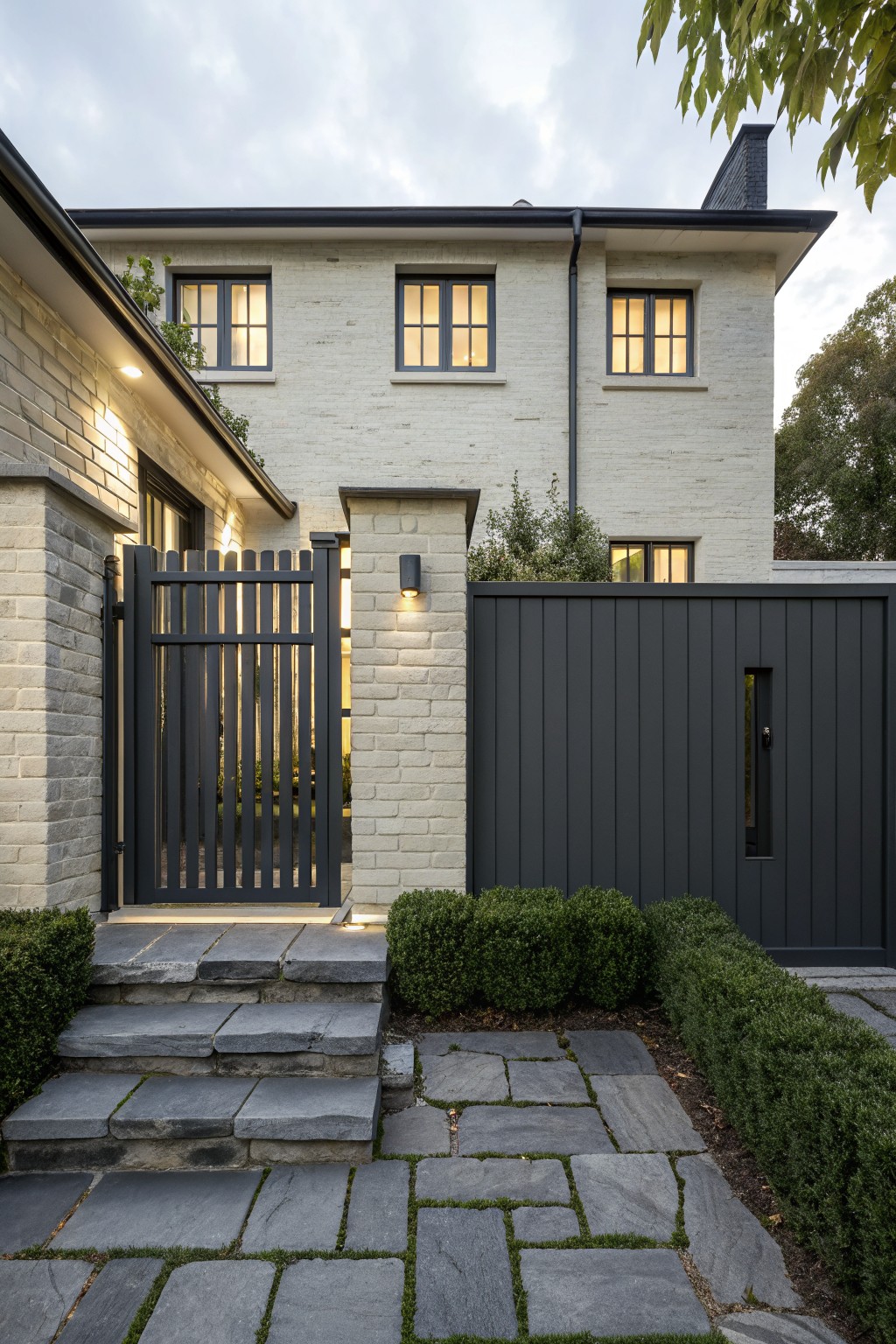 White brick house exterior with black vertical slat metal gate on stone steps, flanked by boxwood shrubs and a tall black slat metal fence panel, shown at dusk with warm lighting.