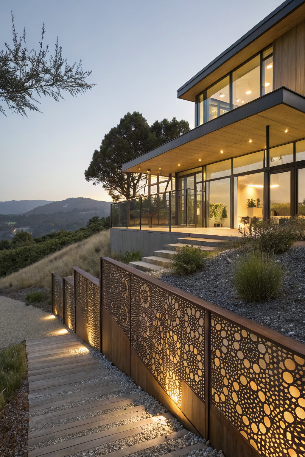 Modern house on a hillside with a wooden pathway lined by illuminated perforated metal screens, steps leading to a balcony with glass railings, large windows, and dry landscaping at dusk.