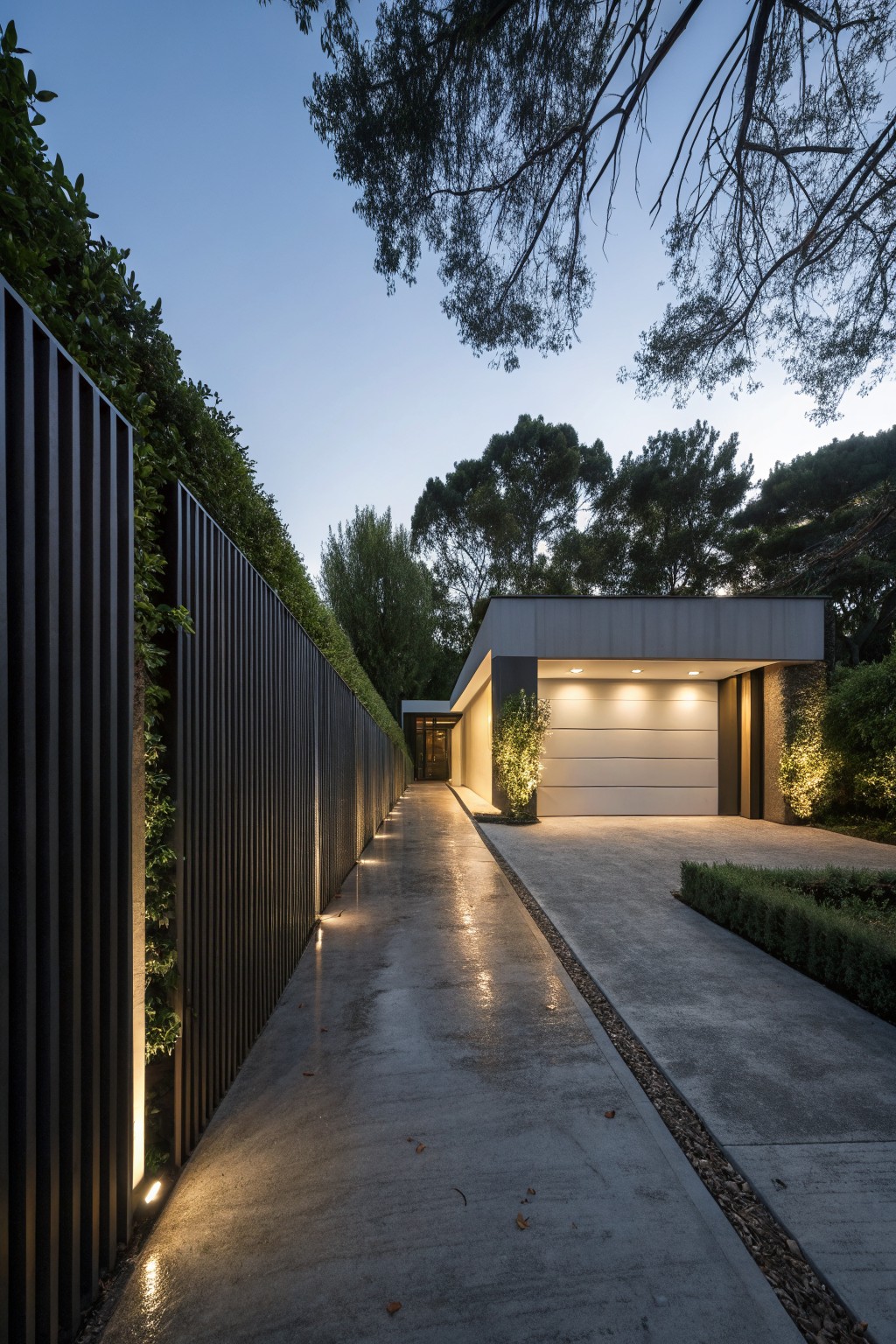 Long wet concrete driveway flanked by tall black slatted metal fence with hedges and base lights, leading to a modern garage with open white door and overhead lights, surrounded by trees at dusk.