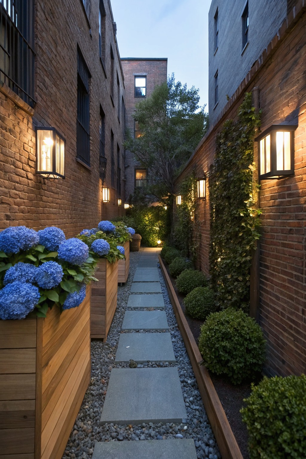 Narrow garden path between tall brick walls lined with wooden planters of blue hydrangeas, stone pavers, gravel edging, boxwood shrubs, ivy on walls, and wall-mounted lanterns at dusk.
